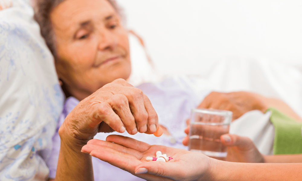 Elderly woman in bed takes pills from a caregiver's hand, holding a glass of water.