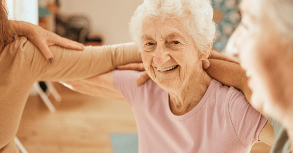Elderly woman smiling during a group exercise class, arms linked with others, wearing a light pink shirt in a bright room.