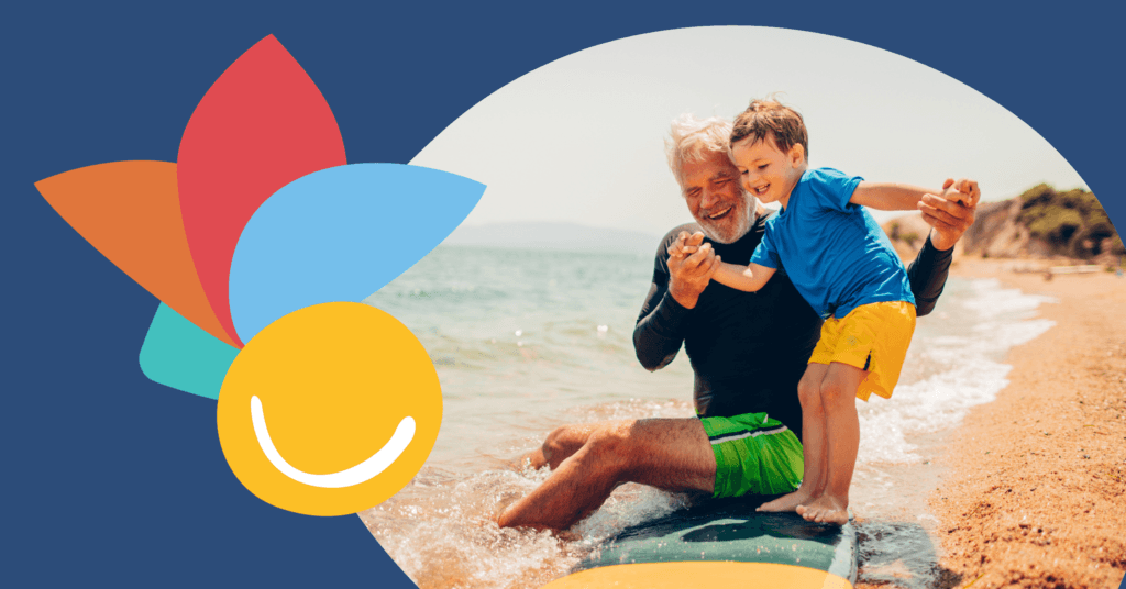 Elderly man and young boy play joyfully on a beach, sitting on a surfboard near the water's edge under a sunny sky.