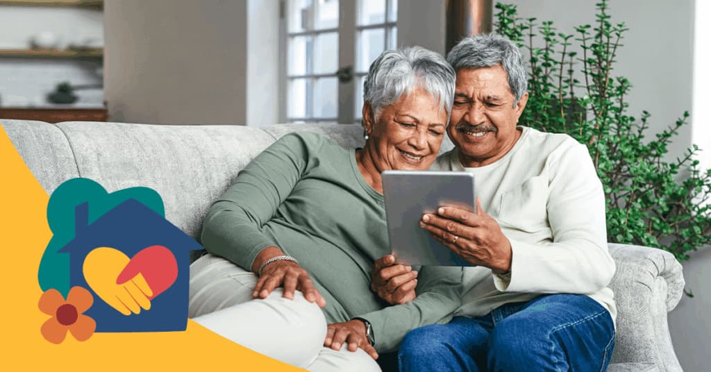 Elderly couple sitting on a sofa, smiling while looking at a tablet. A graphic of a house with a heart and hand is in the foreground.