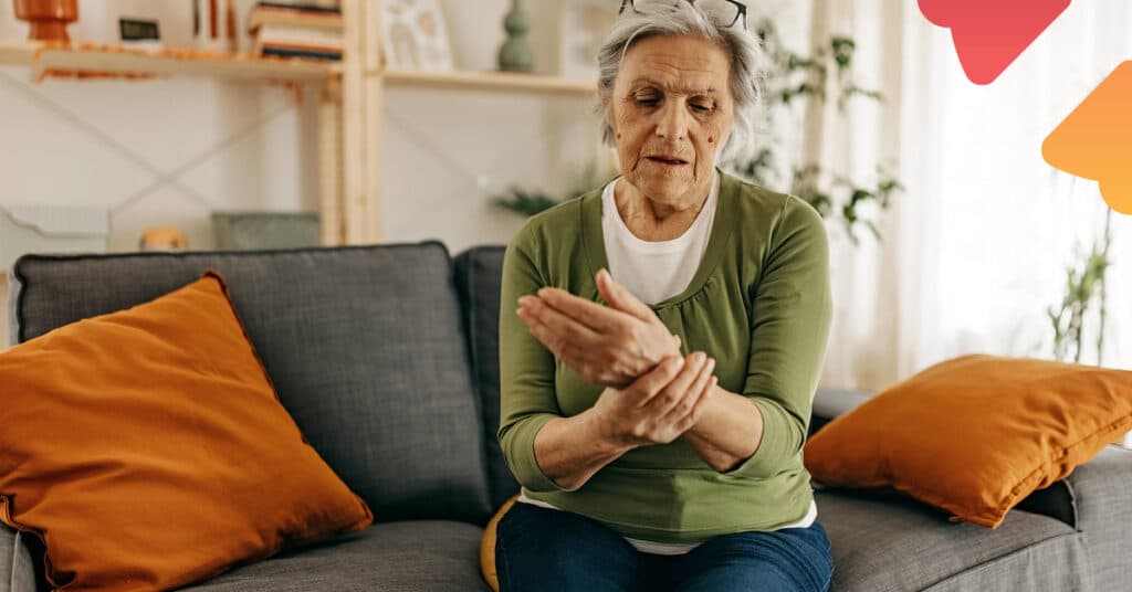 Elderly woman sitting on a couch, holding her wrist with a concerned expression, surrounded by orange cushions in a cozy living room.