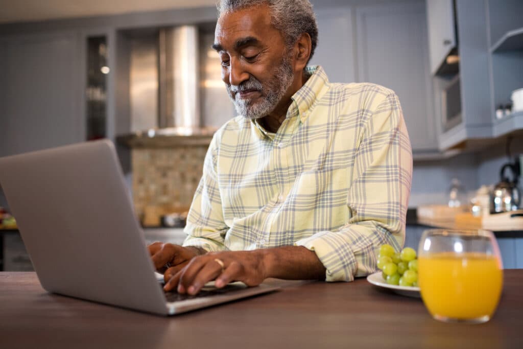 Elderly man in a plaid shirt using a laptop at a kitchen table with a plate of grapes and a glass of orange juice nearby.