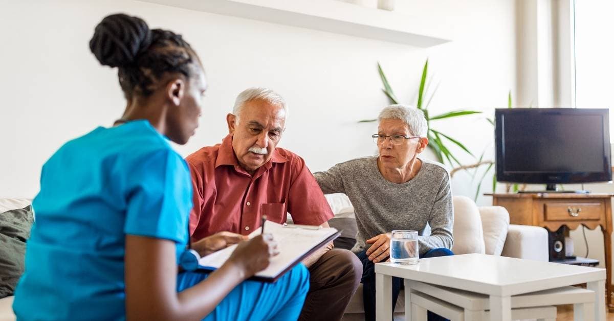 Healthcare worker in blue scrubs discusses paperwork with an older couple sitting on a couch in a living room.