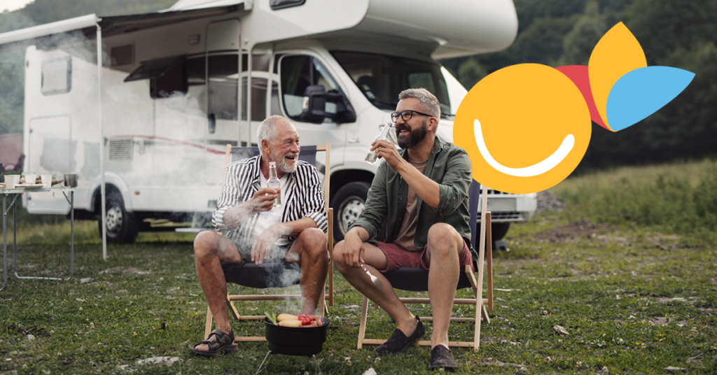 The image shows two men, one older and one younger, sharing a cheerful moment over drinks and a barbecue at a campsite with their motorhome nearby.