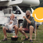 The image shows two men, one older and one younger, sharing a cheerful moment over drinks and a barbecue at a campsite with their motorhome nearby.