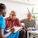 A healthcare professional discusses paperwork with an attentive elderly couple in their home.