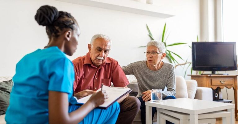 A healthcare professional discusses paperwork with an attentive elderly couple in their home.