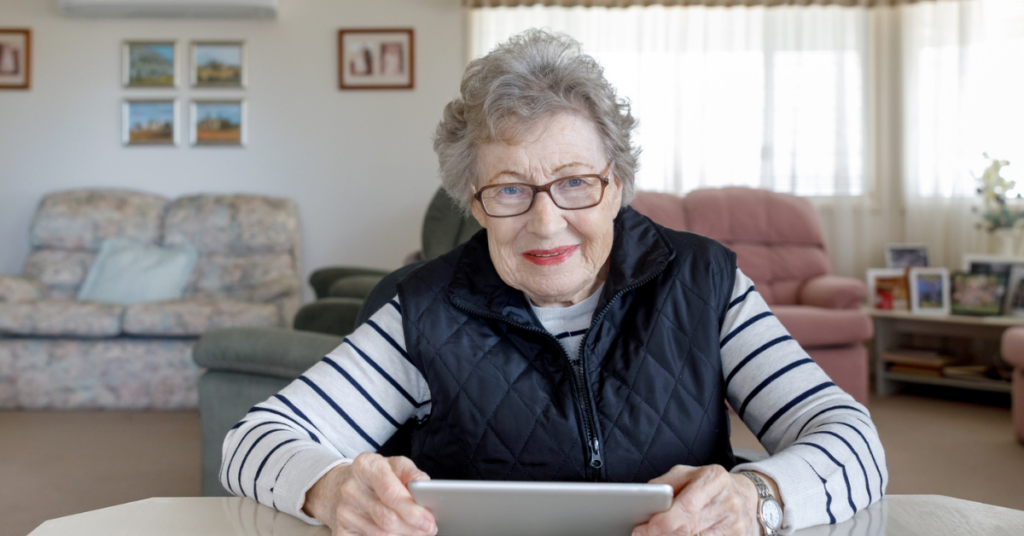 Happy senior lady sitting at table in comfortable lounge room holding an iPad.