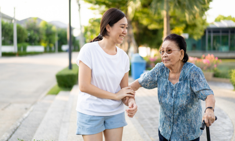 A young woman assists an elderly lady with a cane on a sunny outdoor walk.